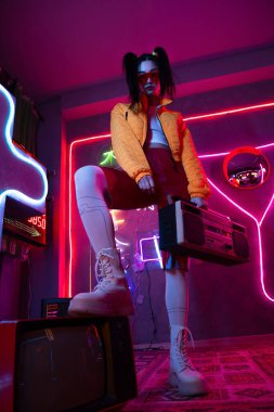 low angle view of young asian woman in sunglasses and orange jacket posing with retro boombox near tv 