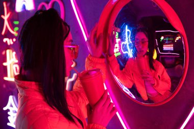 stylish young asian woman in sunglasses posing with paper cup near mirror and neon lighting 