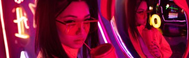 stylish young asian woman in sunglasses posing with paper cup near mirror and neon lighting, banner 