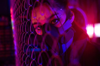 neon lighting on young asian woman in gas mask looking through metallic fence clipart