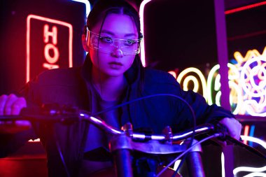 young asian woman in sunglasses riding motorcycle with neon lighting behind 