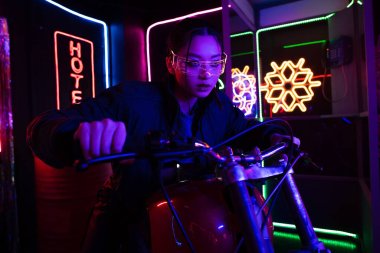 young asian woman in sunglasses riding motorcycle near neon sign outside 