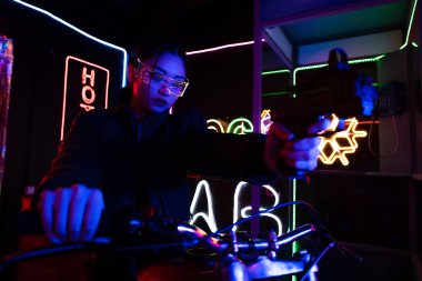 young asian woman in sunglasses aiming with gun while riding motorcycle near neon sign 