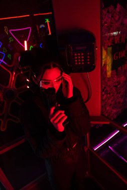 armed asian woman in sunglasses holding gun while talking on retro payphone 