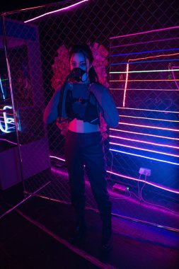 full length of young asian woman in gas mask wearing vest near neon lighting 
