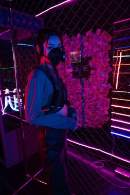 young asian woman in gas mask and headphones standing near neon lighting 