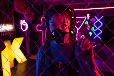 young asian woman in gas mask and wireless headphones looking at camera through metallic fence 