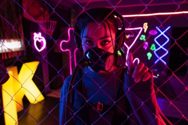 young asian woman in gas mask and wireless headphones looking at camera through metallic fence  clipart