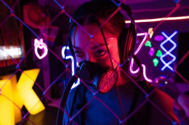 young asian woman in gas mask and wireless headphones near blurred metallic fence 