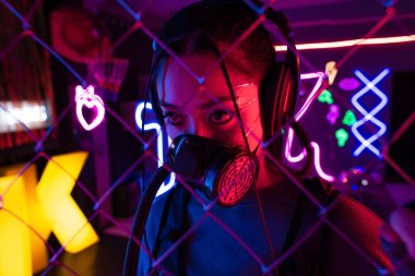 young asian woman in gas mask and wireless headphones near blurred metallic fence  clipart