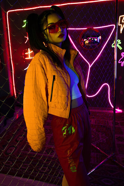 trendy young asian woman in red sunglasses posing near neon sign and metallic fence 