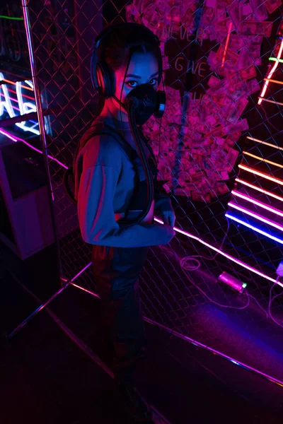high angle view of young asian woman in gas mask and headphones standing near neon lighting 