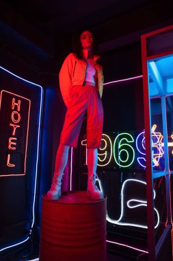 full length of asian woman in sunglasses looking at camera while posing near red neon hotel sign 