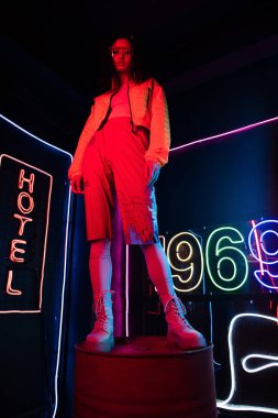 low angle view of asian woman in sunglasses looking at camera while posing near red neon hotel sign 