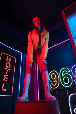 low angle view of asian woman in sunglasses looking at camera while posing near red neon hotel sign 