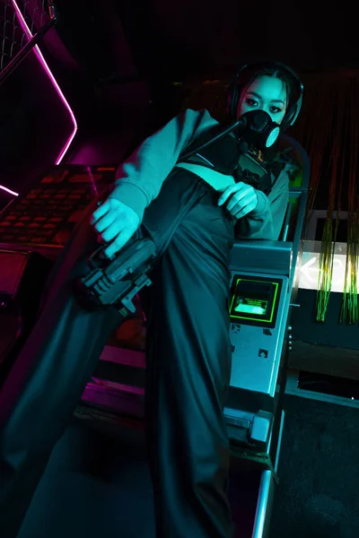 low angle view of armed asian woman in gas mask and wireless headphones looking at camera while holding gun 
