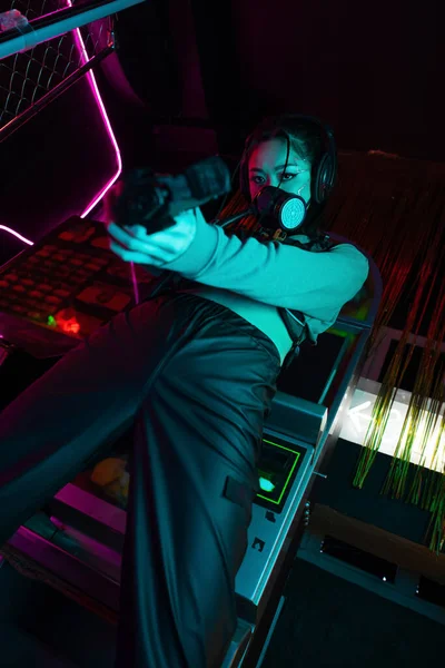 low angle view of armed asian killer in gas mask and wireless headphones aiming with gun 