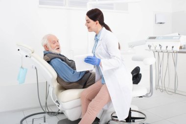 Gülümseyen dişçi diş sağlığı kliniğindeki yaşlı hastaya tablet gösteriyor