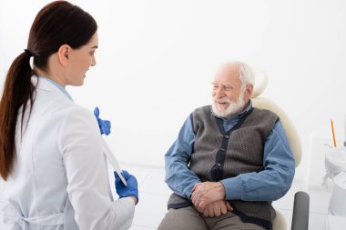Diş hekimindeki kıdemli hasta elinde tabletle gülümseyen doktordan danışmanlık alıyor.