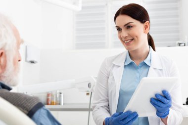 cheerful dentist with tablet in hands looking at senior patient sitting in dental chair clipart