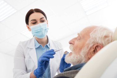 Tıbbi maskeli dişçi, dişçi kliniğinde sonda ve aynayla kıdemli bir adamın dişlerini inceliyor.