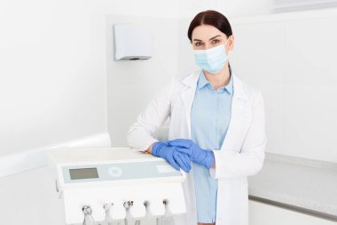 Lateks eldivenli ve tıbbi maskeli dişçi ekipmanın yanında duruyor ve dişçi kliniğindeki kameraya bakıyor.