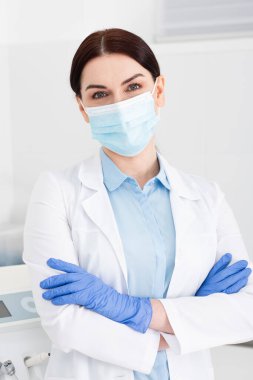 Lateks eldivenli ve tıbbi maskeli dişçi. Diş kliniğinde kolları çapraz duruyor.