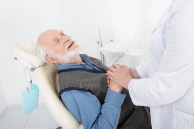 Gülümseyen kıdemli hasta dişçi koltuğunda yatıyor ve klinikte dişçinin elini tutuyor.
