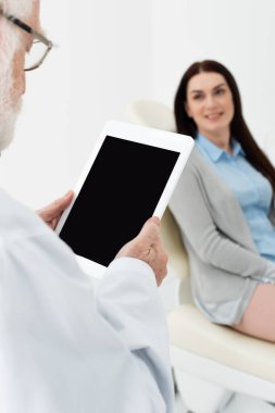Klinikte dişçi koltuğunda bulanık bir kadının önünde boş tabletle duran kıdemli dişçi.