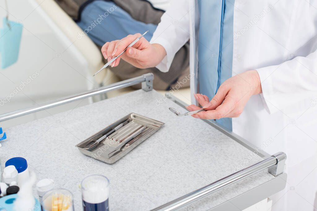 cropped view of female dentist hands holding metal tools above metal tray on table in dental clinic