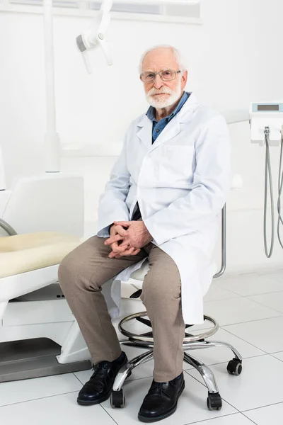 Serious Senior Dentist White Coat Sitting Clenched Hands Dental Clinic — 스톡 사진