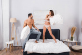 Cheerful woman in lingerie pillow fighting with boyfriend at home 