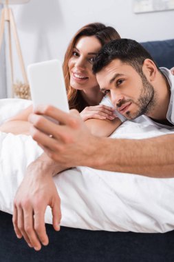 Sakallı adam cep telefonuyla selfie çekiyor. Yatak odasında gülümseyen kız arkadaşının yanında. 