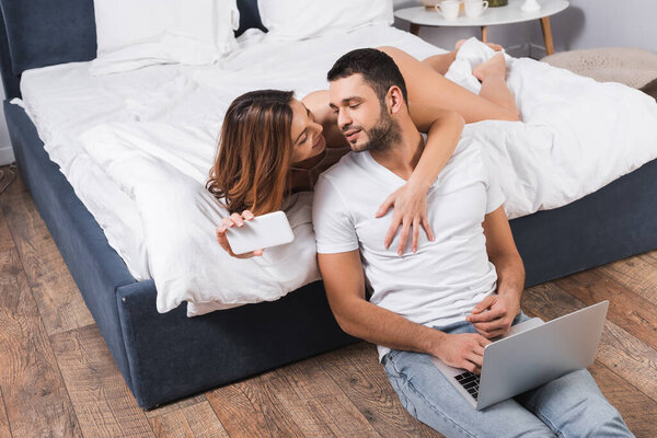 Smiling woman with smartphone hugging boyfriend using laptop in bedroom 