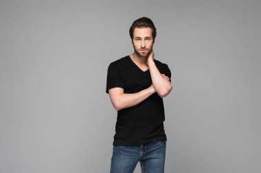 Siyah tişörtlü genç sakallı adam kameraya bakarken gri renkte poz veriyor.