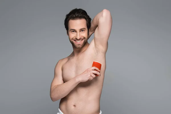 joyful shirtless man applying solid stick deodorant isolated on grey