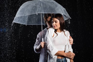 Romantik, genç, yetişkin bir çift şemsiyeyle yağmurda duruyor ve siyah arka planda şehvetle sarılıyorlar.