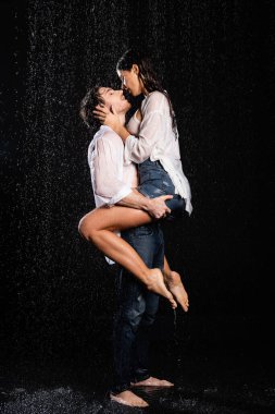 Seksi, beyaz gömlekli ve kot pantolonlu romantik çift siyah arka planda yağmur damlalarında tutkuyla sarılıyorlar.