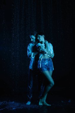 good-looking man gently hugging wet sensual woman in water drops on black background with blue color filter clipart