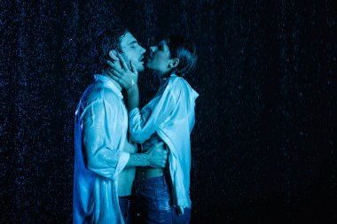 Seksi, ıslak, romantik çift. Mavi filtreli, siyah arka planda su damlalarında nazikçe sarılıp öpüşüyor.