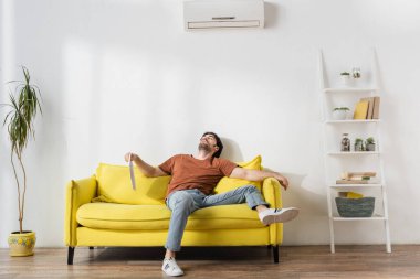 man holding newspaper while lying on yellow couch and suffering from heat in living room clipart
