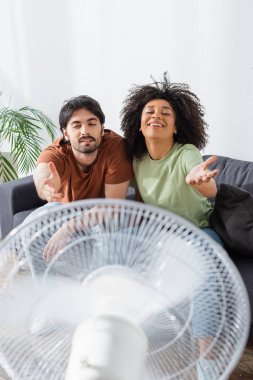 happy interracial couple sitting on couch with outstretched hands near blurred electric fan in living room  clipart