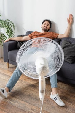 Bulanık elektrikli vantilatörün yanındaki koltukta oturan memnun adam. 