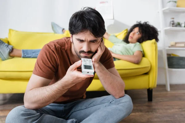 Man Looking Broken Remote Controller Blurred African American Girlfriend Waving — Stock Photo, Image