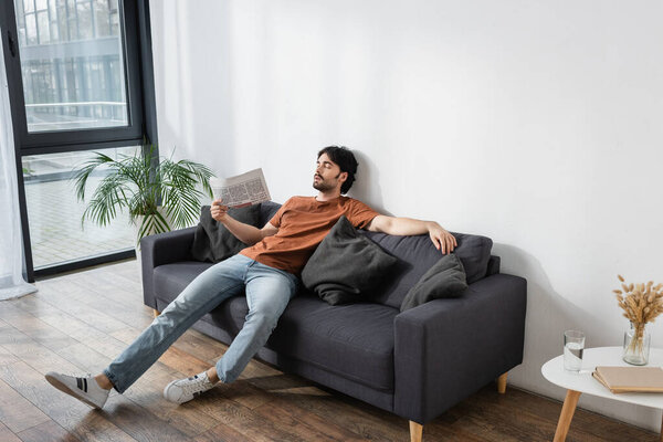 man waving with newspaper while lying on gray sofa and suffering from heat