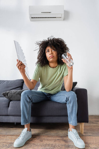 low angle view of african american woman waving with hand fan while holding glass of water and sitting on grey couch