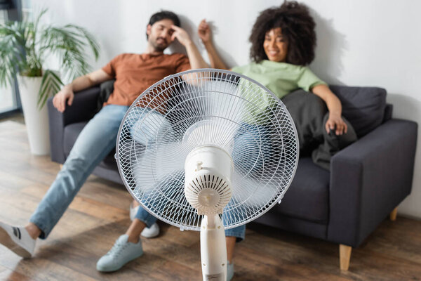 blurred interracial man and woman sitting on couch near electric fan in living room 