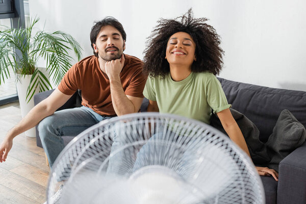 joyful interracial couple sitting on couch near blurred electric fan in living room 