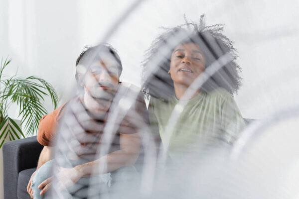 blurred and happy interracial couple near blurred electric fan in living room 