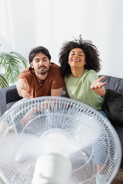 Happy Interracial Couple Sitting Couch Outstretched Hands Blurred Electric Fan — Stock Photo, Image